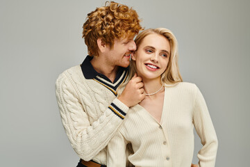 playful redhead man touching neck of joyful blonde woman in trendy clothes on grey, classic fashion