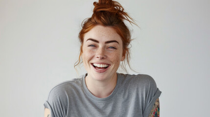 A cheerful woman with red hair tied in a bun, laughing joyfully in a casual grey t-shirt, her...