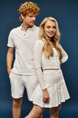 charming blonde woman in white trendy outfit smiling at camera near handsome redhead man on blue