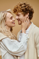 smiling blonde woman hugging face of young redhead man on beige backdrop, old money style concept