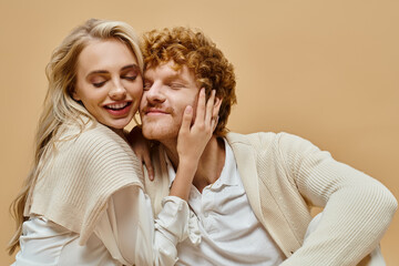 delighted blonde woman with closed eyes embracing head of stylish redhead man on beige backdrop