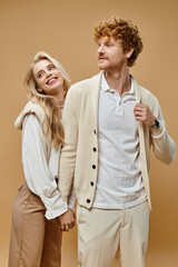 old money style, young cheerful couple in trendy outfit holding hands and looking away on beige