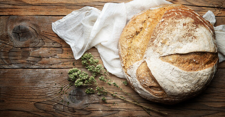 Whole loaf of traditional bread and wild oregano sprig with knife, cutting board and tea towel on wooden background, close-up, copy space.