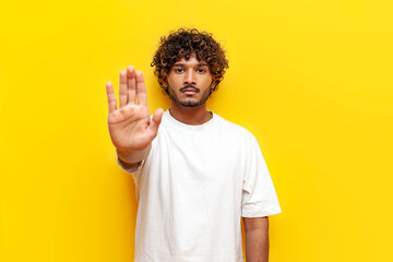 indian man in white t-shirt prohibits and shows stop gesture on yellow isolated background, curly...