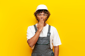 shocked Indian builder in hard hat and overalls covers his mouth with his hand on yellow isolated background, surprised Indian foreman in uniform looks at camera in amazement - obrazy, fototapety, plakaty