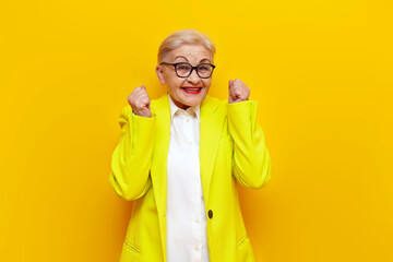 old businesswoman in formal wear wins and rejoices at success on yellow isolated background, elderly pensioner grandmother in blazer celebrates victory and good luck