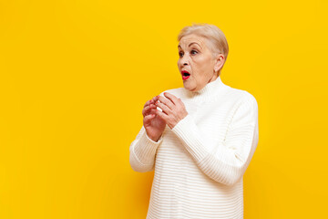 frightened old grandmother in white sweater is surprised and worried on yellow isolated background,...