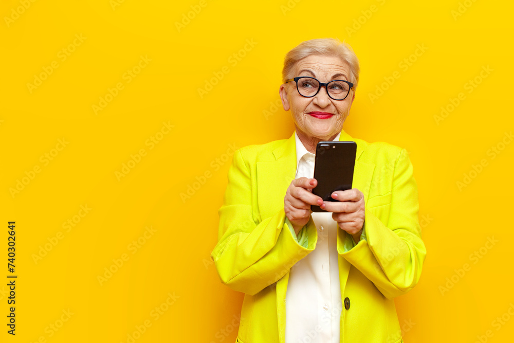 Wall mural pensive old businesswoman in formal wear using smartphone on yellow isolated background, elderly pen