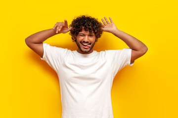 curly indian man makes faces and makes a fool of himself showing his tongue on a yellow isolated background, a young guy makes fun of and shows foolish grimaces - obrazy, fototapety, plakaty