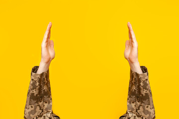 hands of Ukrainian soldier in camouflage military uniform raised up holding and showing empty space on yellow isolated background, empty military hands advertising copy space - obrazy, fototapety, plakaty