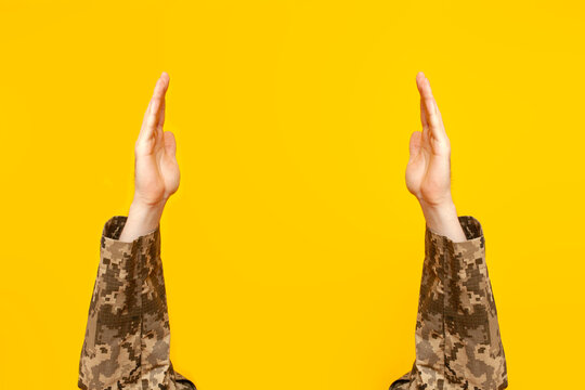 hands of Ukrainian soldier in camouflage military uniform raised up holding and showing empty space on yellow isolated background, empty military hands advertising copy space