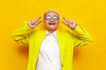 crazy old businesswoman in glasses and formal wear showing peace gesture and tongue out on yellow...