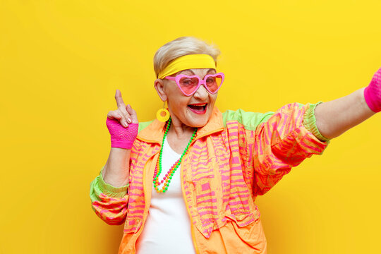 crazy funny old granny in sports colorful clothes takes a selfie on a smartphone and screams on a yellow isolated background, an elderly woman in youth hipster clothes takes a photo of herself