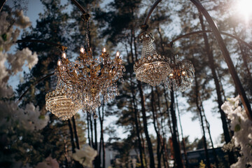 Luxurious crystal chandeliers in the middle of a pine forest in the glow of the sunset. Fabulous wedding decor with chandeliers.