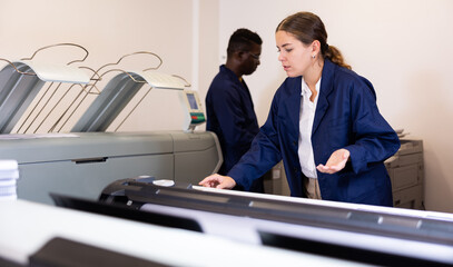 Concentrated young female typographer in a blue robe uniform pressing the buttons on the plotter in...