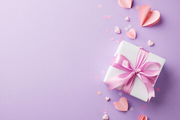 Obraz na płótnie Canvas Graceful offerings: the art of giving to women. Top view shot of white gift box, pink satin ribbon, paper hearts, confetti on lavender background with space for seasonal greetings or promo content