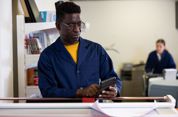 Attentive middle-aged African American male specialist in a blue uniform using calculator in the printing house