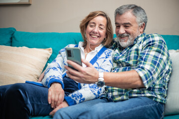 Retired couple laughing while making a video call with cell phone on sofa