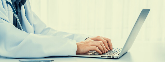 Doctor at hospital sit at his desk working on laptop diagnosing patient test results, developing...