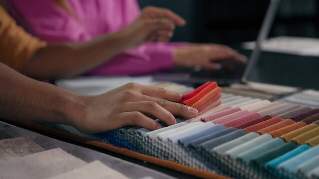Close up details woman assistant and man designer choosing color fabric samples hands touch textile swatches velvet upholstery material for furniture home interior design project on laptop at workshop