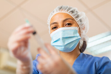 Positive woman healthcare worker wearing disposable medical cap and face mask holding syringe for injection or vaccination.