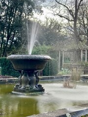 Water fountain in an English Garden with plants and a pergola in the background.  Taken in Battersea Park, London UK