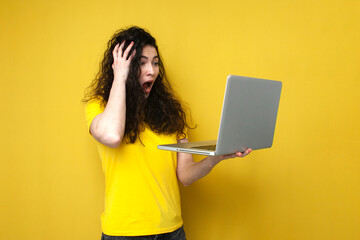 shocked curly girl in yellow t-shirt holds laptop and shows surprise by opening her mouth on yellow...