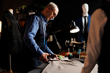 Precise experienced elderly suitmaker manufacturing suit in tailoring studio workspace, cutting fabric material with scissors. Process of manufacturing upcoming bespoke fashion design collection