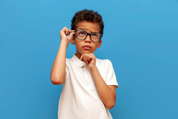 african american puzzled teenager boy in white polo and glasses thinks and plans on blue isolated...