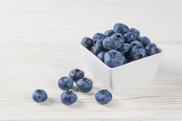 Fresh blueberries in a small square white bowl with scattering of berries on white wooden background. Organic berries, healthy food, wild berries