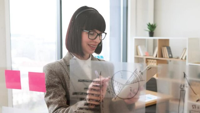Concentrated caucasian woman in headphones writing on transparent board with sticky papers in conference hall. Serious young female worker rewriting notes from tablet in meeting room.
