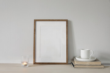 Blank vertical wooden picture frame mock up on wooden table. Modern interior. Cup of coffee on pile of vintage books, burning candle. White wall background. Scandinavian home. Poster display template.