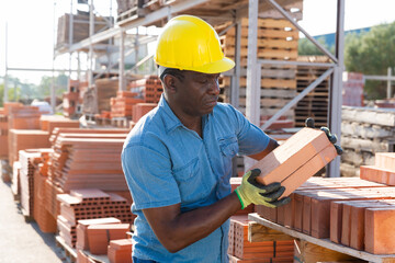 African-american male worker carrying clay bricks in outdoor construction material storage.