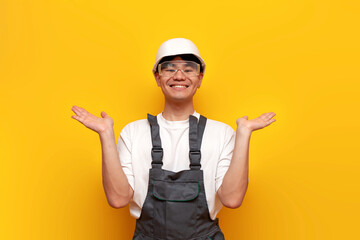 asian male builder in uniform holding empty hands on yellow isolated background