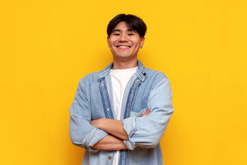 asian young man in denim shirt standing with arms crossed over yellow isolated background