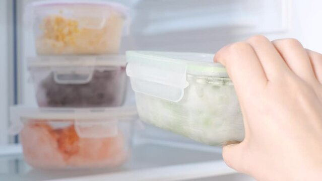 Female hand holds a glass container with green peas against the background of freezer with other boxes.