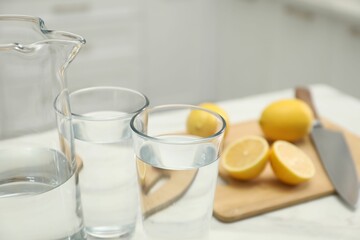 Jug, glasses with clear water and lemons on white table in kitchen, closeup