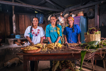 A beautiful portrait of three beautiful Latin women, with many ears of corn on the table, they smile in front of the camera.