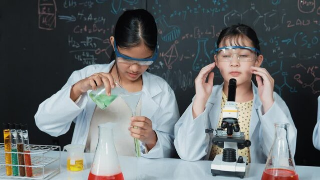 Young high school student doing experiment by pouring sample in test tube. Girl wearing glasses and looking under microscope to inspect sample in STEM science class. Creative education. Edification