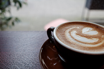 A freshly brewed latte with a heart-shaped art on the foam, sitting on a dark brown ceramic saucer...
