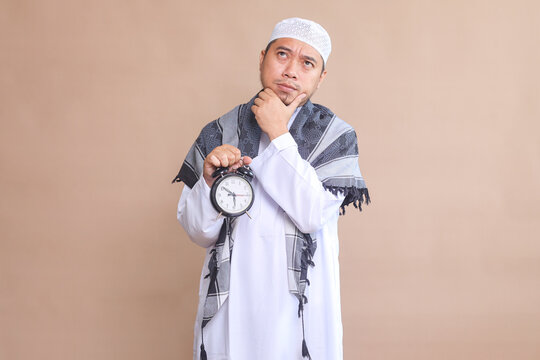 Young Asian Man Wearing Muslim Robe Gesture Thingking While Holds Alarm Clock, Waiting for Iftar Time at Ramadan Month