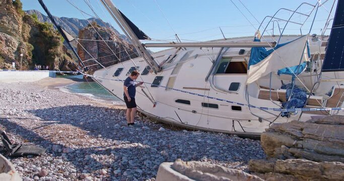 A curious passerby examines and photographs site of the wreck of sailing ship fenced off with protective film. A man films on smartphone sunken yacht washed up on beach, seashore crime scene, accident