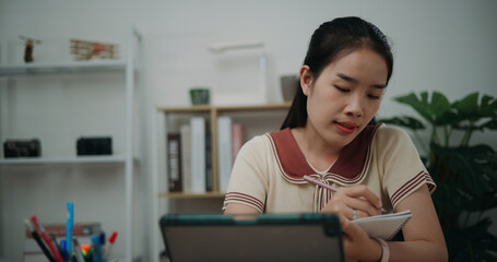 Selective focus, Front view, Female writer sitting at desk thinking idea holding pen making notes in diary while working on tablet for online learning at home, creative thoughts to journaling