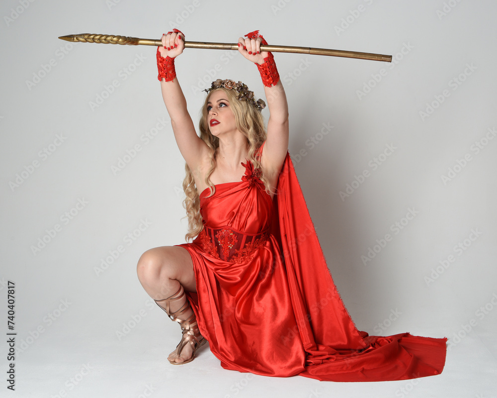 Wall mural Full length portrait of beautiful blonde model dressed as ancient mythological fantasy goddess in flowing red silk toga gown, crown. kneeling pose,  golden trident weapon, isolated studio background