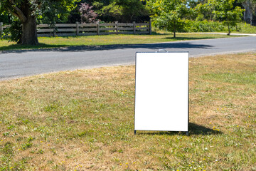 Blank white background texture of a standing board sign on the grass in a rural setting. Empty mockup template of an advertisement panel to display information or attract customers in the countryside.