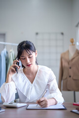 Young asian merchant women using smartphone to talking about customer information and clothes orders to taking notes while working and shipping delivery for online shopping business in home office