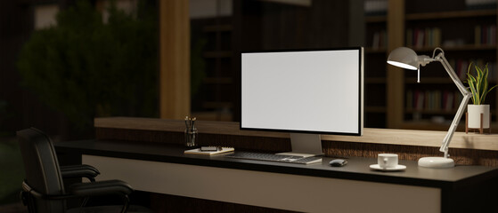 An office desk with a computer mockup, a table lamp, and decor in a modern dark office room at night