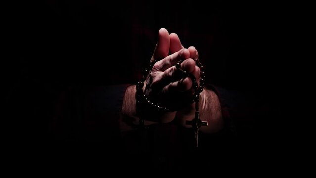 A man's hands wrapped with a wooden Rosary comes out of the dark into the light with praying hands to Jesus.