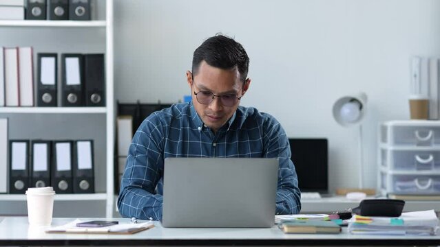 Technology concept, dark-skinned Asian businessman wearing glasses sits and works with a laptop computer. Stack of documents and cell phones for contacting work within the office.