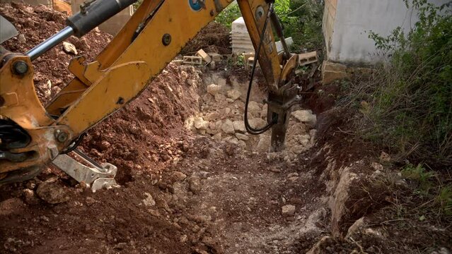 Slow motion of a backhoe loader with an hydraulic hammer attached drilling breaking rocks in a hole for a pool